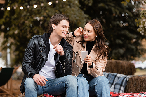 cheerful young woman holding smartphone while listening music and sharing wired earphones with boyfriend