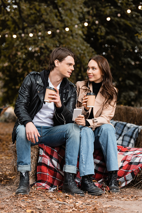 cheerful young woman and man holding paper cups while listening music and sharing wired earphones