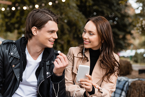 cheerful young woman holding smartphone while listening music and sharing wired earphones with happy boyfriend