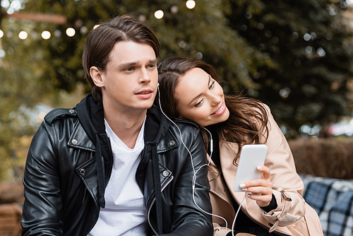 cheerful woman holding smartphone while sharing wired earphones with boyfriend and listening music together