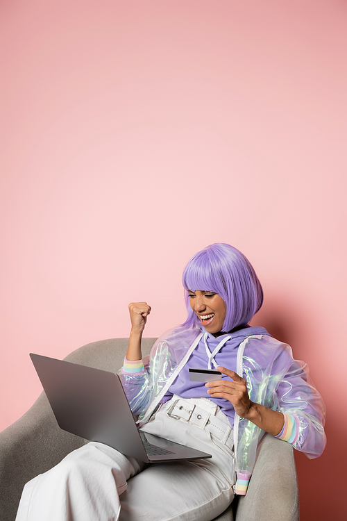 excited african american woman in purple wig using laptop while holding credit card and doing online shopping on black friday on pink