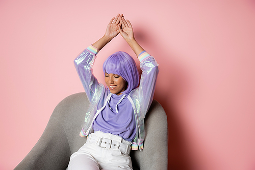cheerful african american woman in purple wig sitting with raised hands on grey armchair on pink