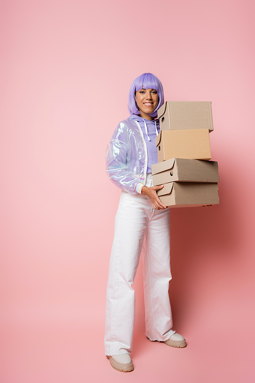 full length of happy african american woman in purple wig holding carton boxes on pink