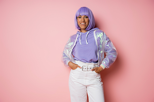cheerful african american woman with purple hair posing with hands in pockets on pink
