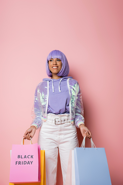 joyful african american woman with purple hair holding shopping bags with black friday lettering on pink