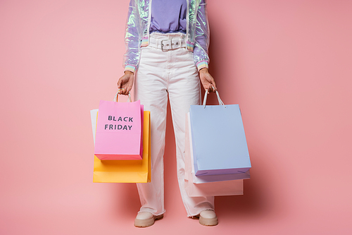 cropped view of woman in transparent jacket and white pants holding shopping bags with black friday lettering on pink