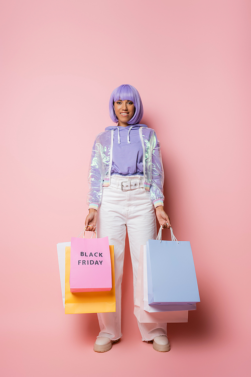 cheerful african american woman with purple hair holding shopping bags with black friday lettering on pink