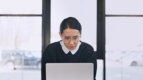 serious and thoughtful businesswoman in eyeglasses looking at laptop in office