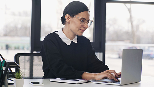 smiling businesswoman in eyeglasses and formal wear typing on laptop near smartphone