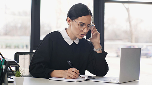 brunette businesswoman in eyeglasses talking on smartphone near laptop and writing in notebook