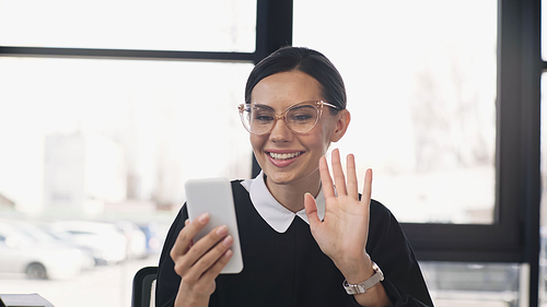 cheerful businesswoman in eyeglasses waving hand during video call on smartphone