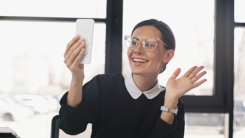 excited businesswoman in eyeglasses waving hand at mobile phone during video chat