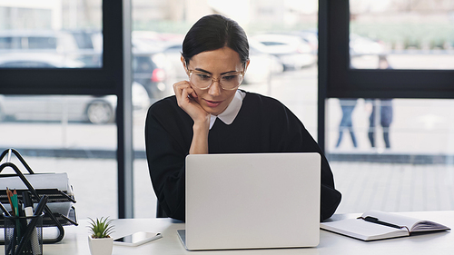 thoughtful businesswoman in eyeglasses sitting with hand near face at laptop