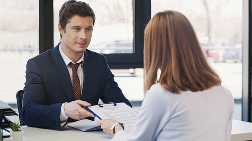 positive man in formal wear giving clipboard with resume to businesswoman on job interview