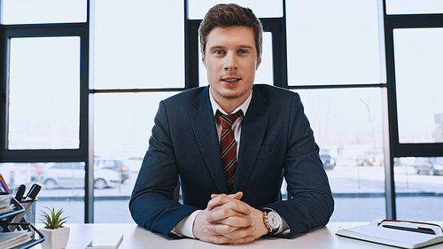positive businessman sitting at workplace with clenched hands and looking at camera
