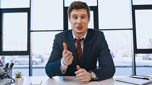 positive businessman pointing with hand and looking at camera at workplace in office