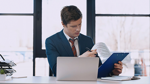 serious businessman looking at document near laptop in office