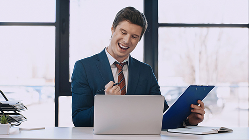 excited businessman holding folder and showing win gesture near laptop