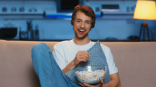 happy young man eating popcorn while watching movie in living room