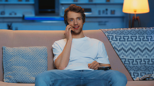 happy young man holding remote controller while talking on smartphone in living room