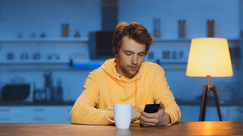 focused young man using smartphone near cup of tea on wooden table