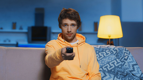 young man in yellow hoodie sticking out tongue while clicking channels in living room