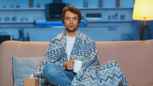 young sick man sitting under blanket and holding cup of tea in living room