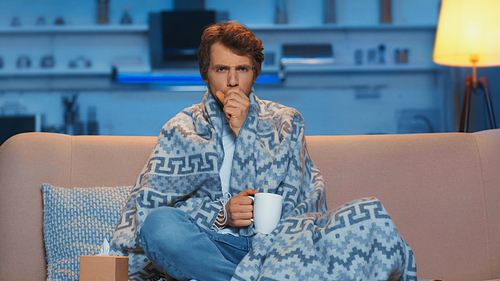 young sick man sitting under blanket and coughing while holding cup of tea in living room