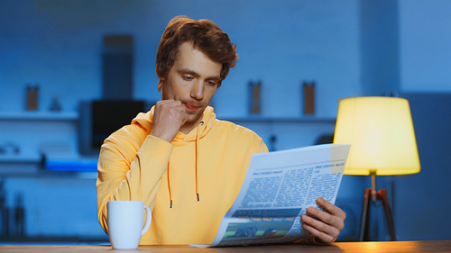 young man in yellow hoodie reading newspaper near cup of tea at home
