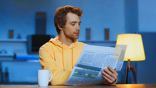 young man in yellow hoodie reading newspaper near cup with beverage at home