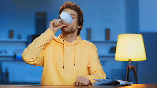 young man in yellow hoodie drinking tea near newspaper on table