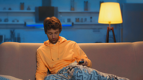 sleepy young man in yellow hoodie holding remote controller and sitting on sofa