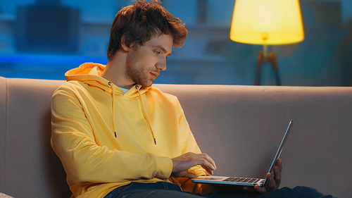 young freelancer in yellow hoodie using laptop while working from home in evening