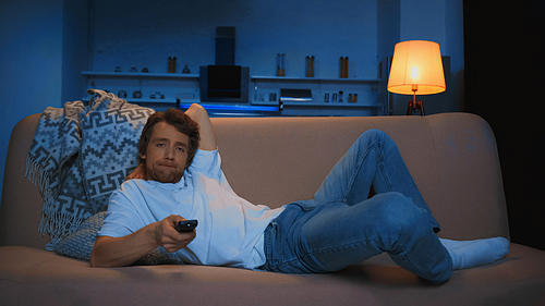 bored young man lying on couch and watching tv in modern living room at night
