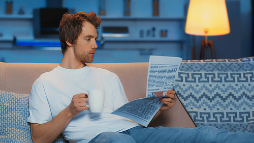 young man in white t-shirt reading newspaper and holding cup of tea while resting on couch
