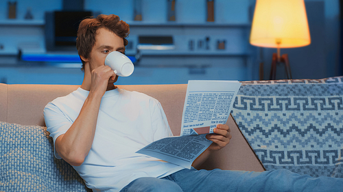 young man in white t-shirt reading newspaper and drinking tea while resting on couch