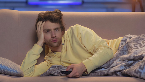 sleepy young man in yellow hoodie resting under blanket and holding remote controller at night