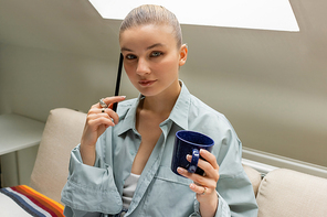 Young woman holding pen and cup while looking at camera on couch in living room