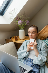 Young woman holding smartphone and looking at laptop and sitting on couch in living room