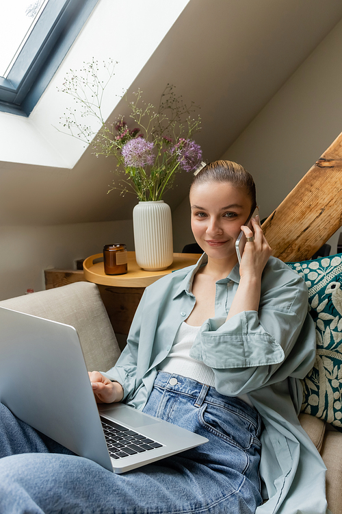 Smiling freelancer talking on smartphone and looking at camera near laptop at home