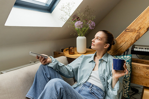 Young woman holding cup and smartphone while sitting on couch at home