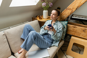 Young woman in earphone holding cup near laptop on couch at home