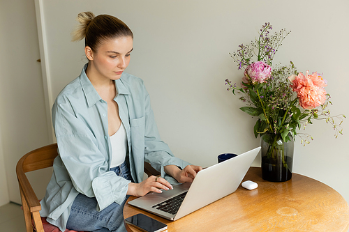 Young woman using laptop near cup and flowers at home