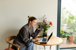 Young freelancer using laptop and holding cup near flowers at home