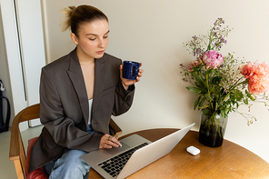 Young woman in blazer using laptop and holding cup near bouquet on table at home
