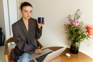 Woman in jacket holding cup and looking at camera near laptop and flowers at home