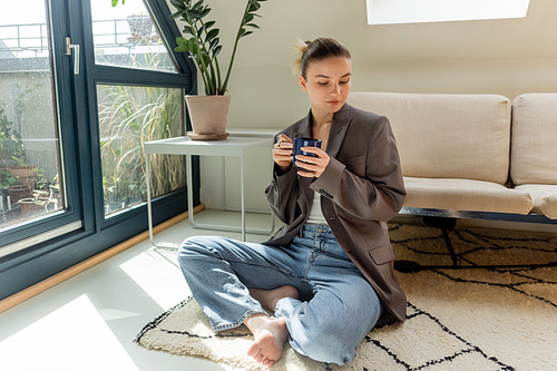Woman in jacket holding cup while sitting on carpet in living room