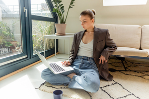 Young freelancer in jacket using laptop near cup on carpet at home