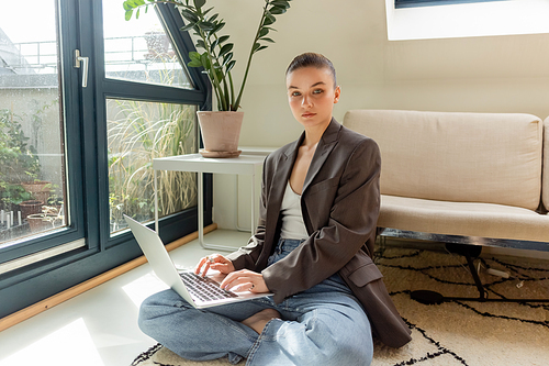Young woman in blazer using laptop and looking at camera on carpet at home
