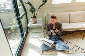 Freelancer in blazer holding cup and looking at laptop on carpet at home